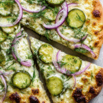 Close-up of a pickle pizza topped with sliced pickles, red onions, fresh dill, and melted cheese on a golden-brown crust.