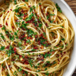 Close-up of spaghetti coated in garlic-infused olive oil, garnished with parsley, red pepper flakes, and grated cheese.