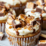 Close-up of a mini cheesecake topped with chopped Snickers candy, caramel drizzle, and chocolate sauce.